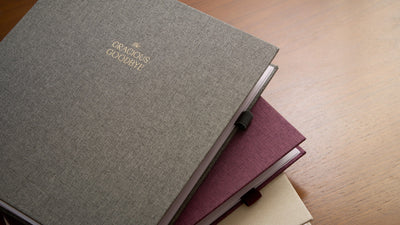 Three linen planners stacked on a warm wooden table, the gray one on top with its gold title.