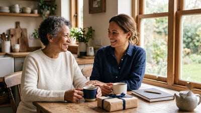 A mother and her adult daughter talking over tea at a sunlit kitchen table, a wrapped gift set to one side.