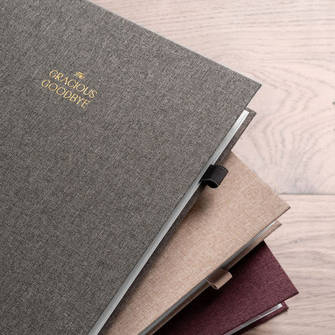 The Gracious Goodbye end-of-life planner in gray, merlot, and sanddune linen, stacked closed with gold-foil covers.