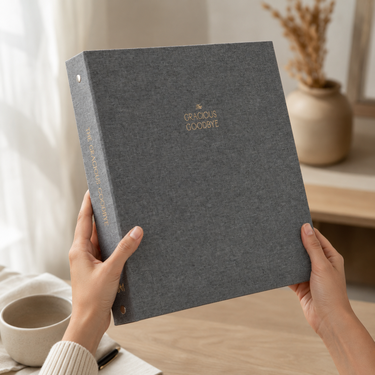 Hands holding the closed gray linen Gracious Goodbye legacy binder, showing its large 12-inch format.