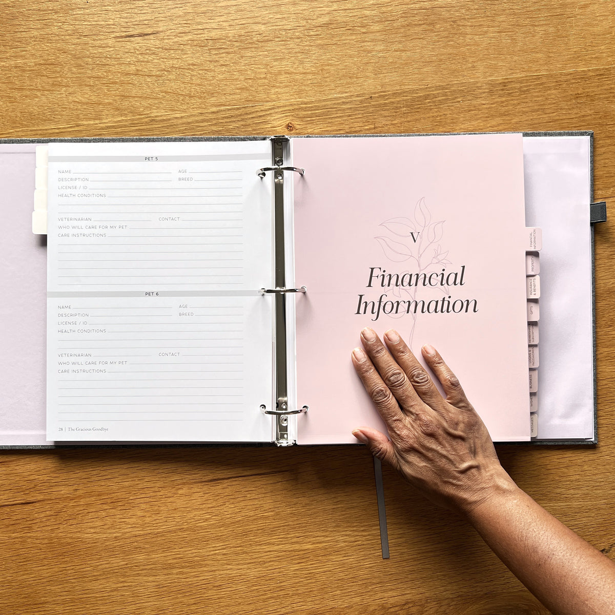 A hand resting on the Financial Information divider of The Gracious Goodbye planner, open on a wooden table.