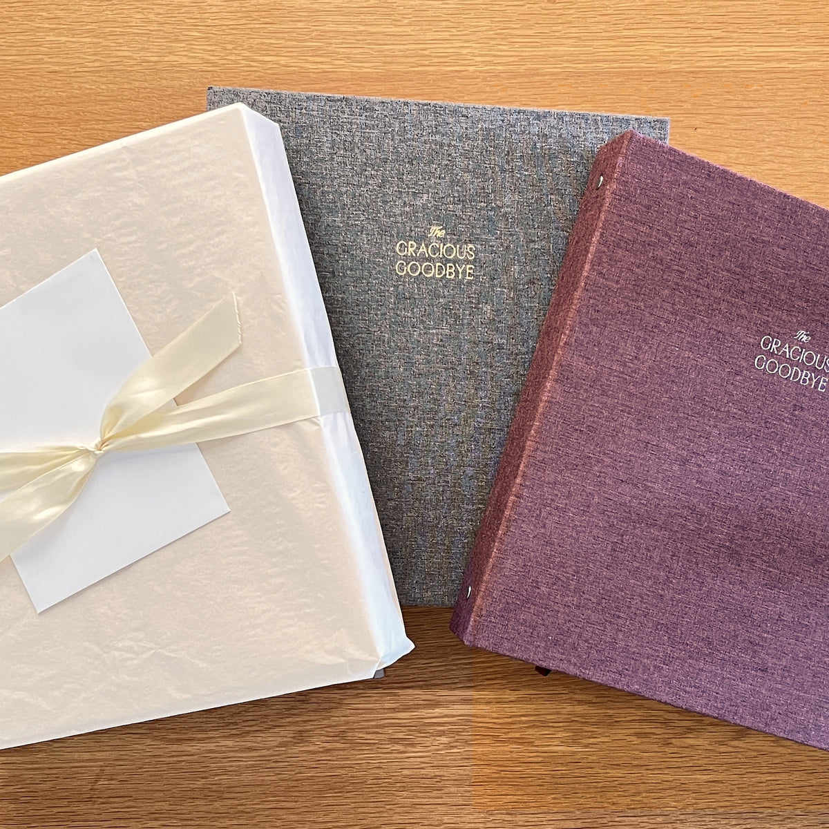 A gift-wrapped planner with a cream ribbon and blank card beside The Gracious Goodbye planners in gray and merlot linen on a wooden table.
