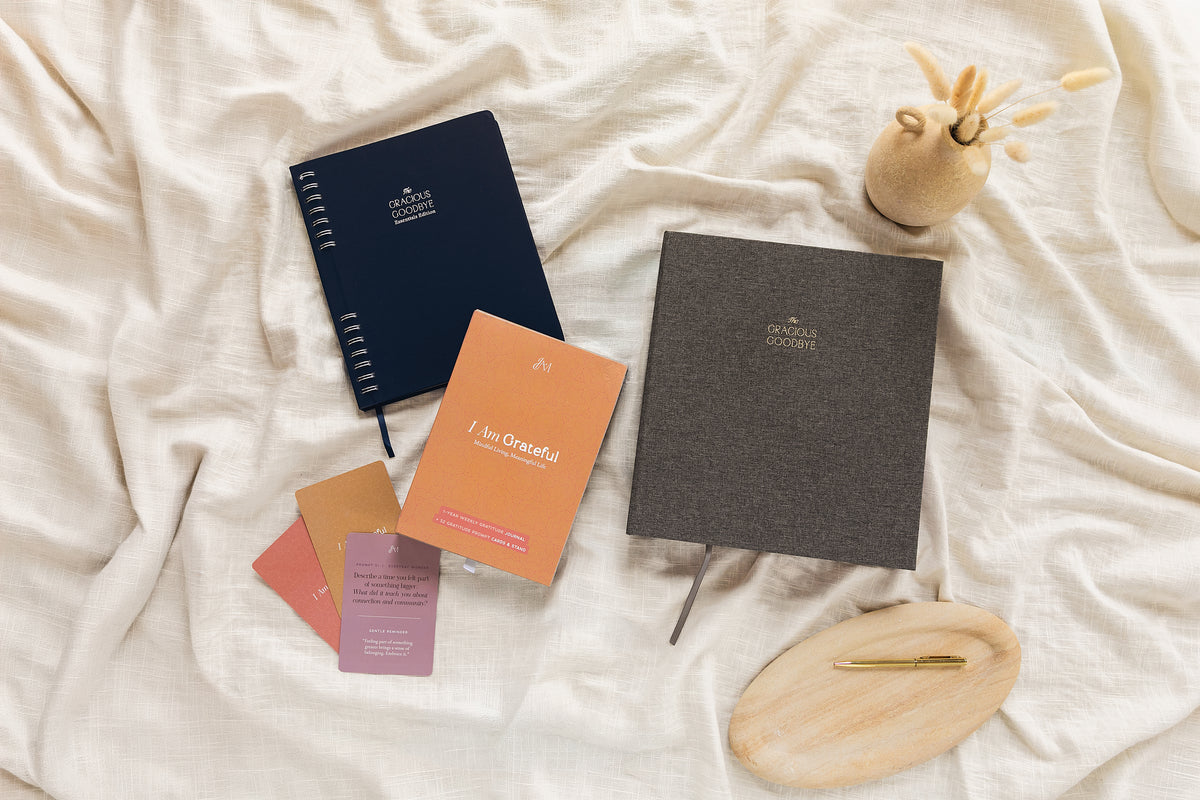 The Gracious Goodbye planner in grey linen, The Gracious Goodbye Essentials, and the I Am Grateful gratitude journal set with its prompt cards, arranged on cream linen with a gold pen.